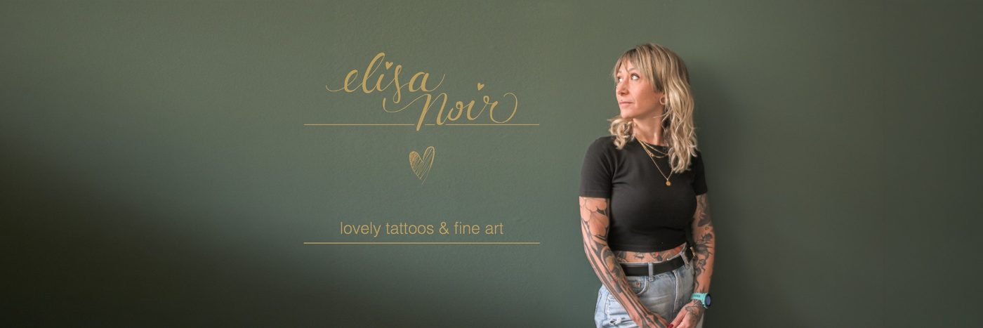 Elisa Noir, Tätowiererin im Herzstickl, vor einer grünen Wand mit dem Schriftzug „elisa noir, lovely tattoos & fine art“